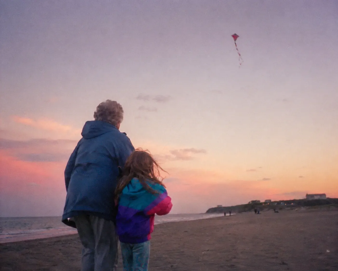A child and grandparent on a beach at dusk, looking up at a kite